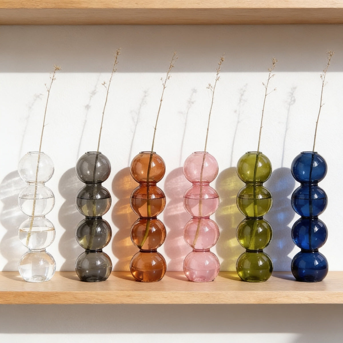 Seven stacked glass bubble vases in a row on an oak shelf, one of each colour.