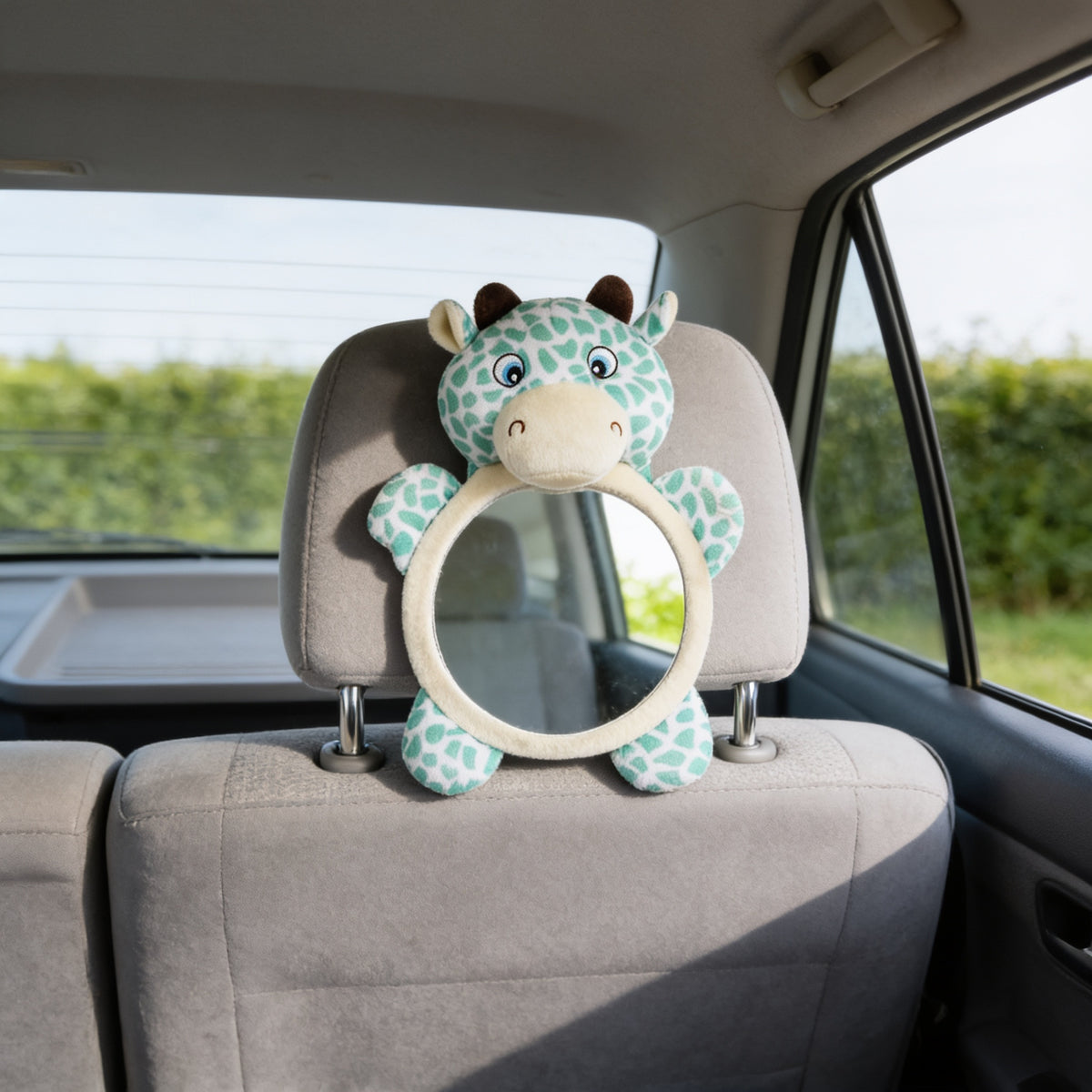 Deer design rear-facing baby car mirror with mint spotted plush around a round mirror, strapped to an empty grey back-seat headrest with a sunlit hedge through the rear window