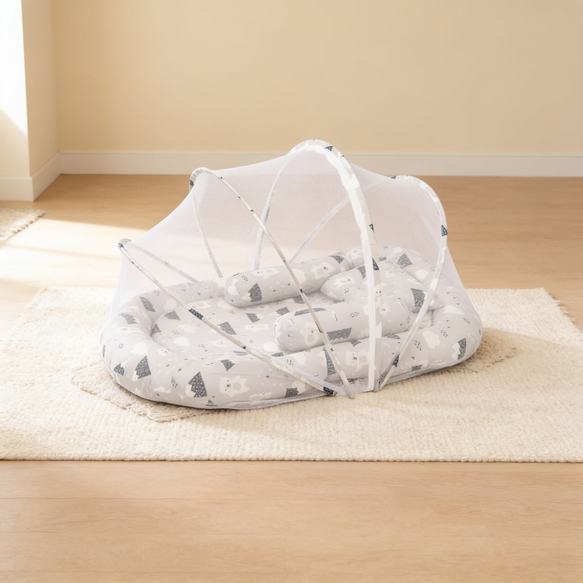 Grey Mountains foldable travel bassinet, white cotton with grey mountain and cloud shapes, with its mesh net dome up on a cream rug in a bright living room