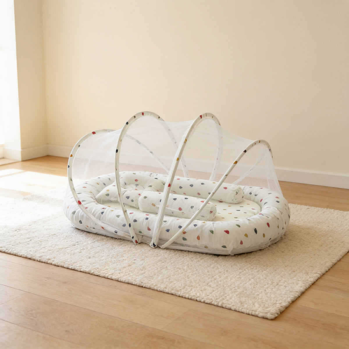 Sage Dots foldable travel bassinet, white cotton with small dark teardrop dots, with its mesh net dome up on a cream rug in a bright living room