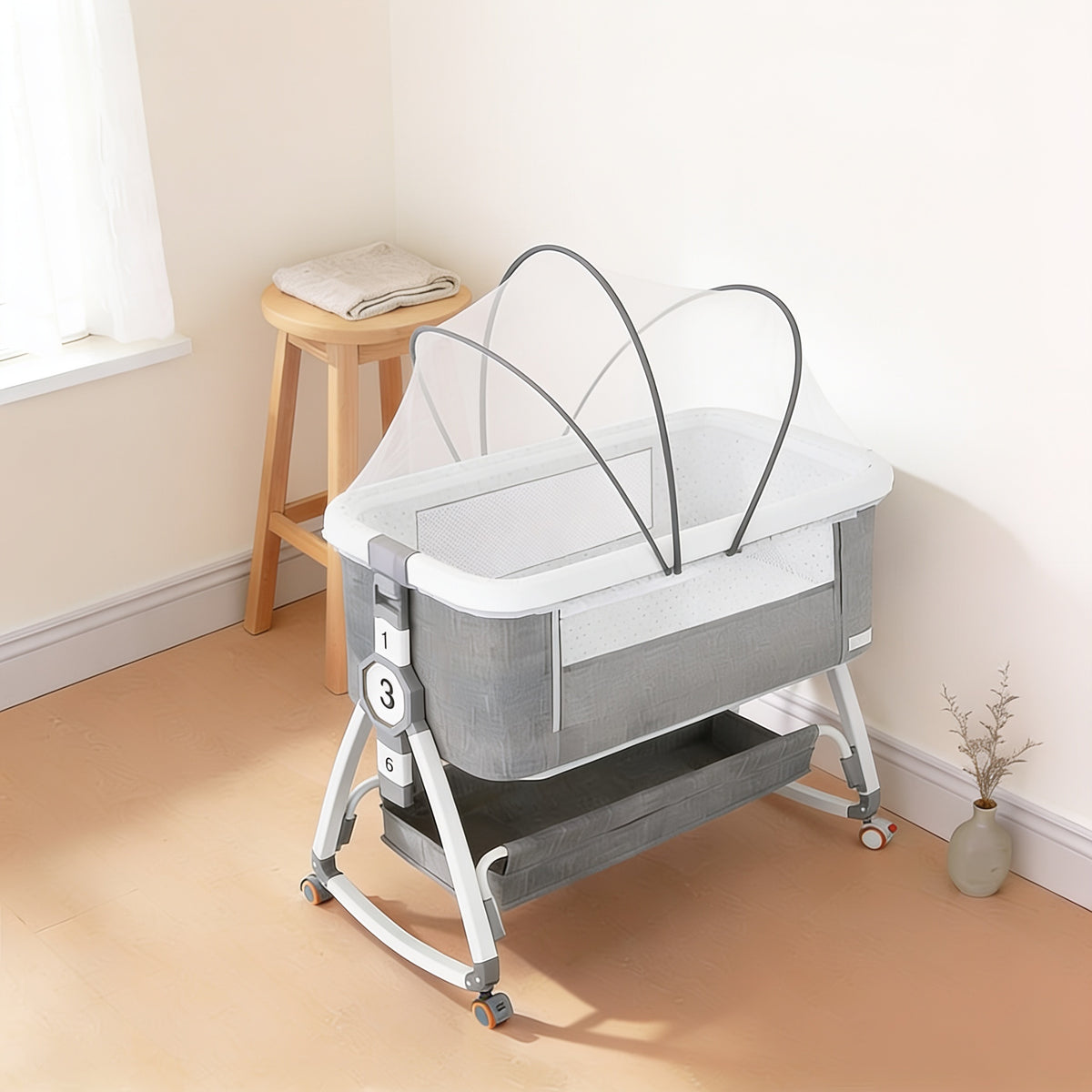 Grey and white bedside bassinet in a cream nursery corner with its sheer white mosquito net fitted over the top on thin grey folding ribs, two tall arches crossing above the empty mattress and a rounded lobe over each end, grey storage basket below