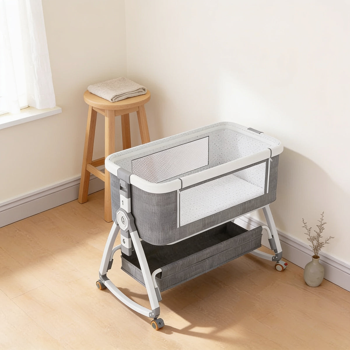 Grey and white bedside bassinet fully closed in a cream nursery corner, its white mesh ventilation panel facing the camera, shown beside a tall oak stool with a folded oatmeal blanket and a painted window sill for size
