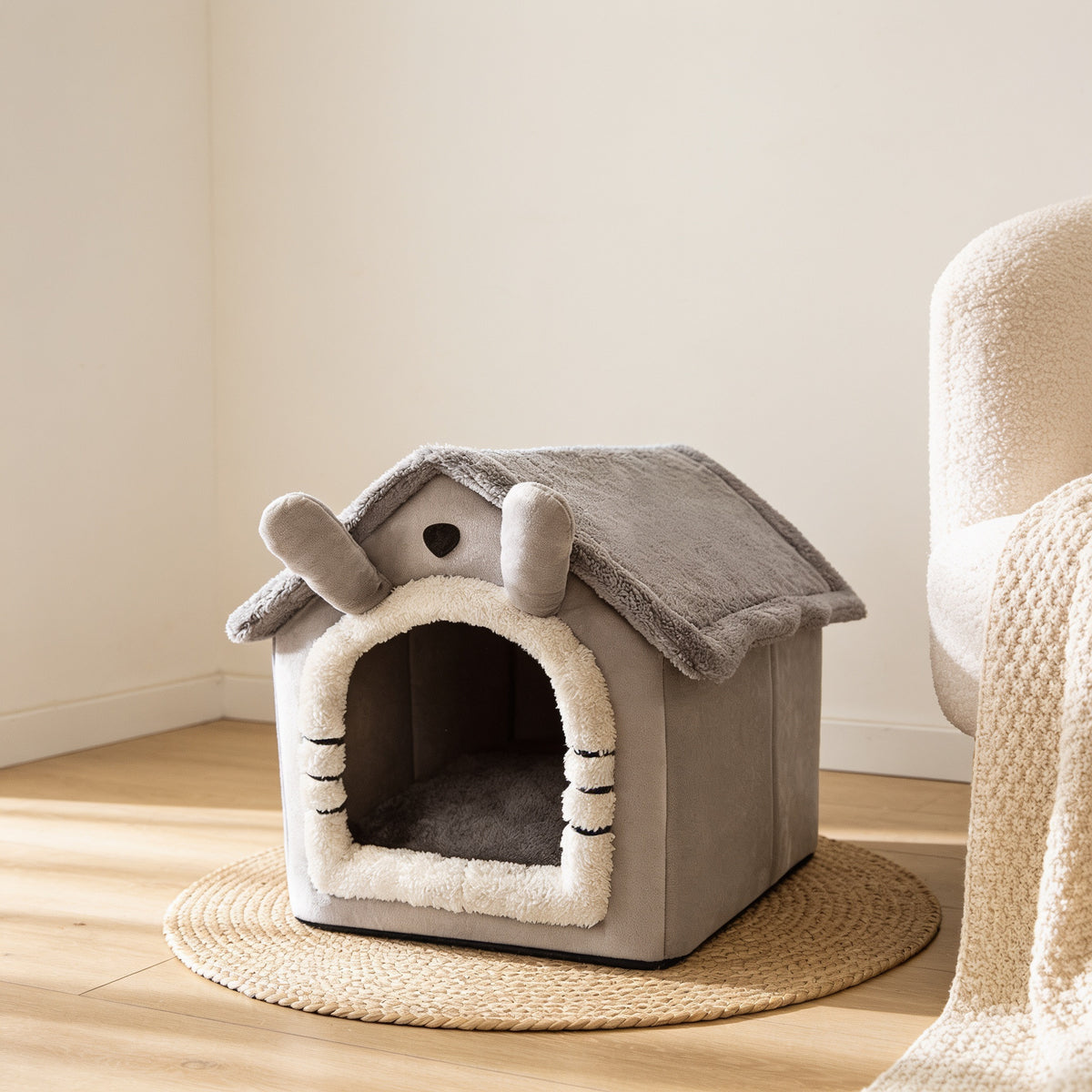 Gray house-shaped covered cat and dog bed with cream trim around the doorway on a jute rug in a cosy corner