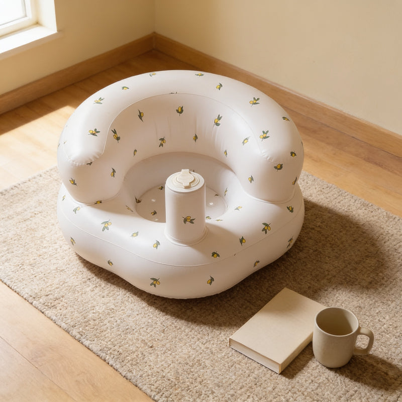 Olive Flower inflatable baby floor seat, empty, on an oatmeal woven rug beside a plain cream book and a stoneware mug in a warm cream living room corner lit from the window on the left