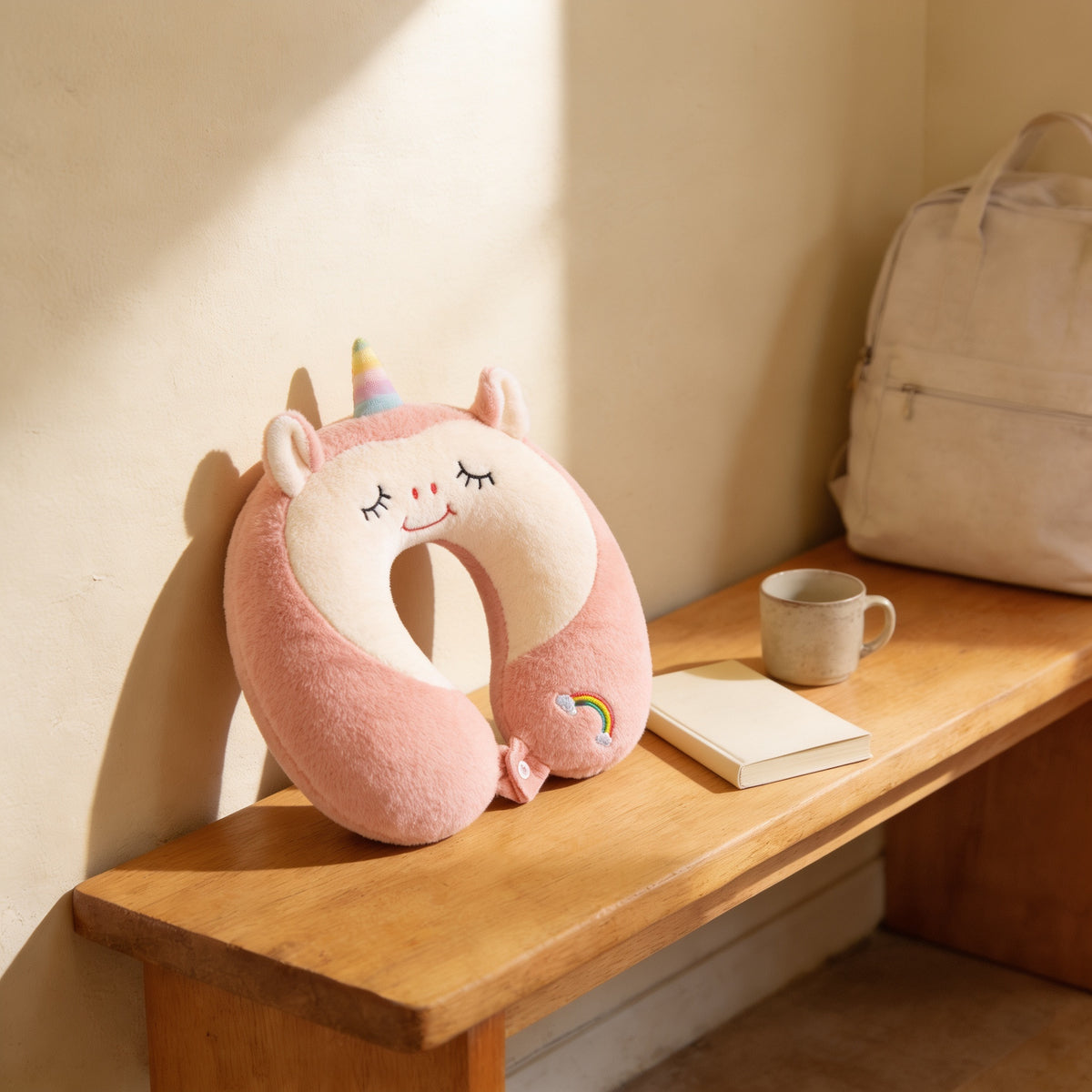 Pink unicorn memory foam travel neck pillow resting on a light oak entryway bench beside a packed canvas bag