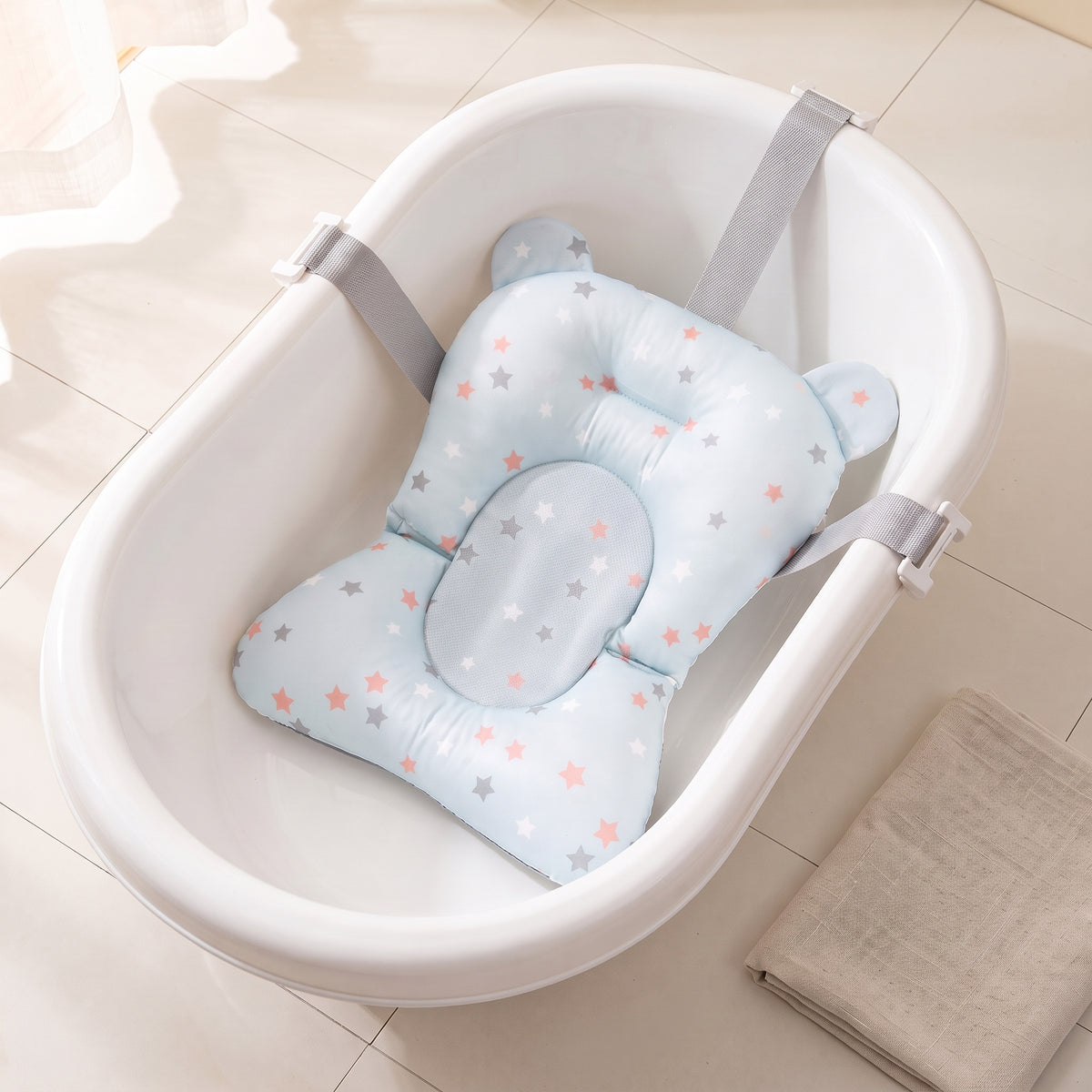 Blue Stars non-slip newborn baby bath cushion fitted in an empty white baby bathtub, its grey belts clipped over the rim, on a cream tiled bathroom floor beside a folded oatmeal towel