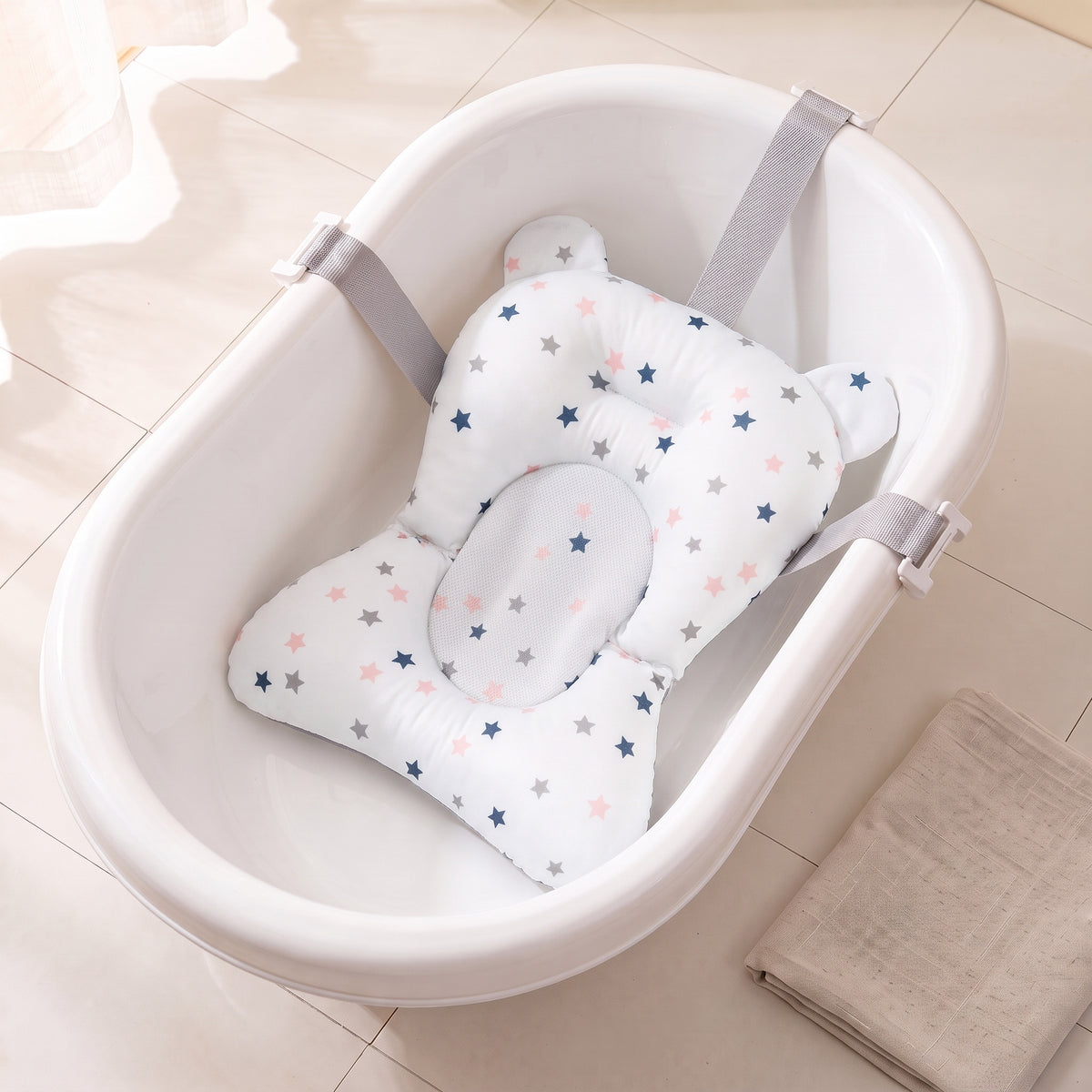 White Stars non-slip newborn baby bath cushion printed with navy, grey and pink stars, fitted in the same empty white baby bathtub with its belts clipped over the rim