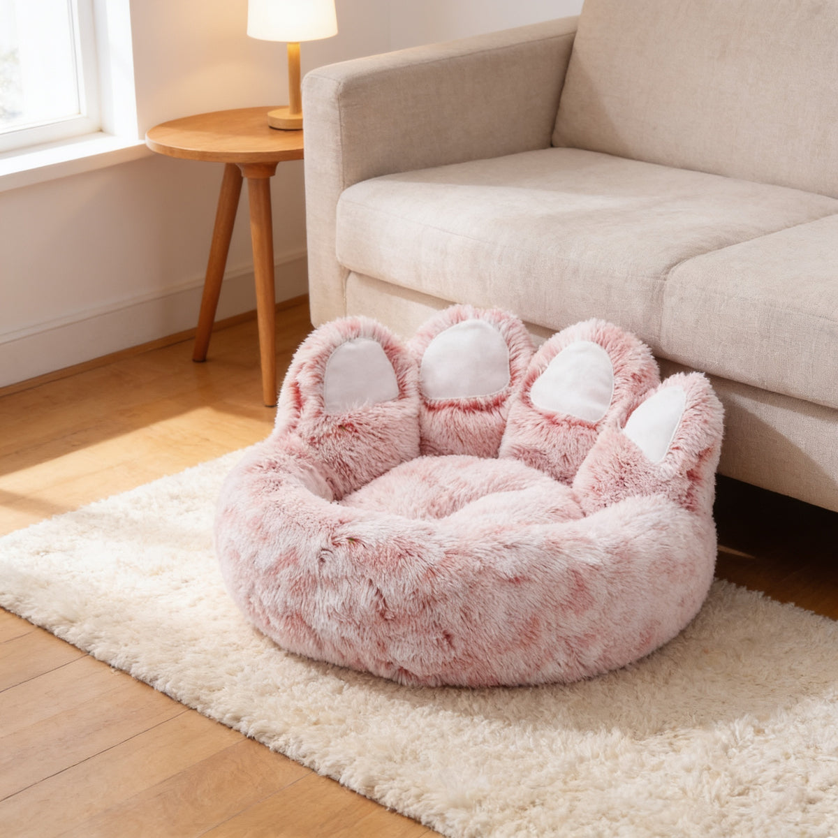 Pink paw-shaped plush dog and cat bed with white toe pads on a cream rug beside a linen sofa