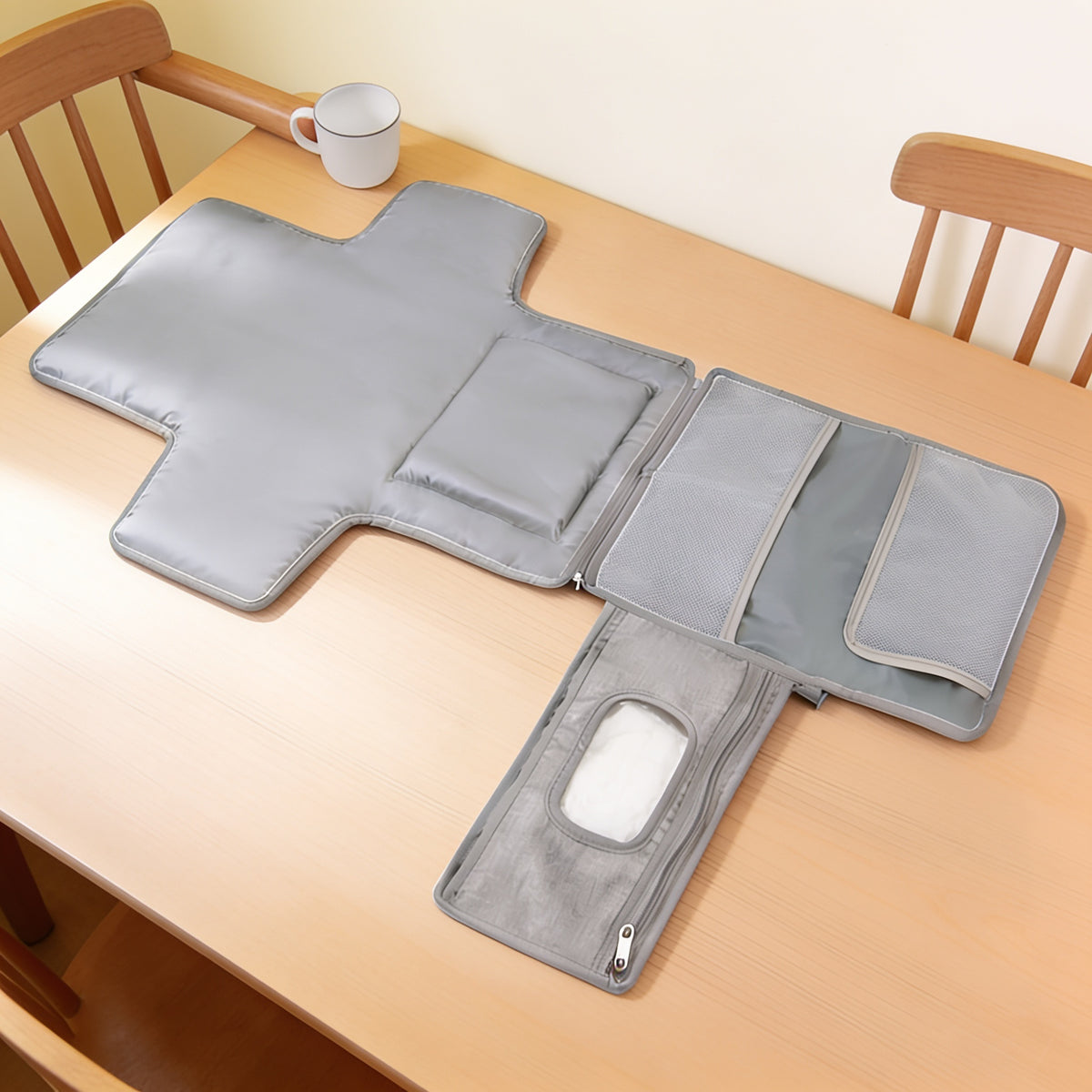 Grey portable diaper changing pad opened flat on a light oak dining table beside a plain white mug: cross-shaped padded mat with a raised padded panel, zip-joined flap with two empty mesh pockets, and a wipes panel with plain wipes behind its clear window