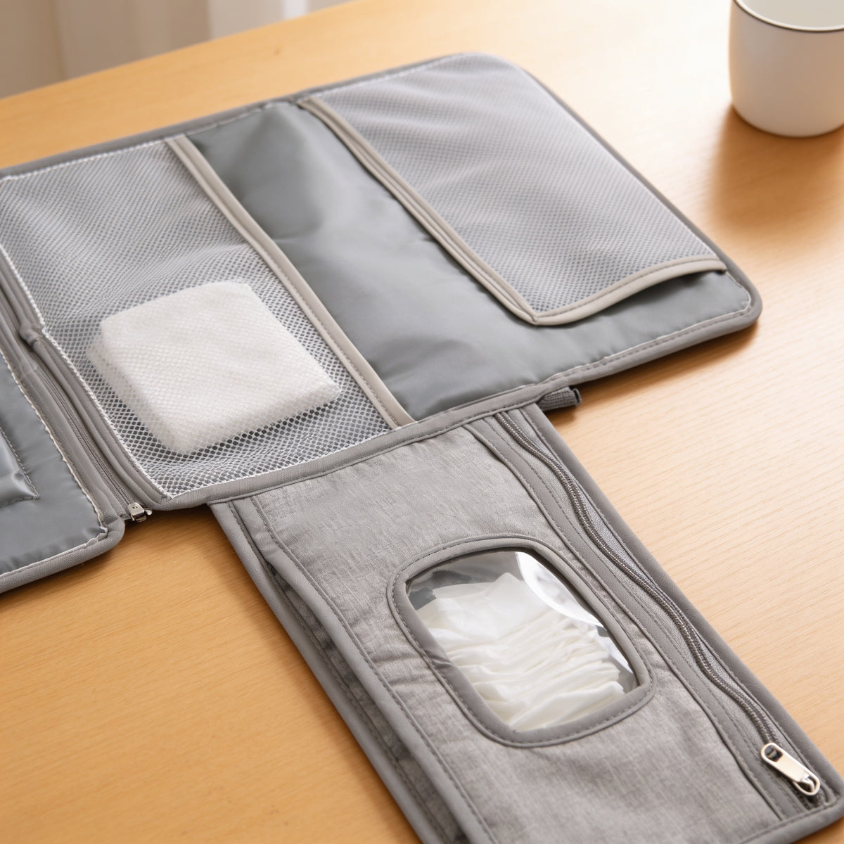 Close-up of the grey changing pad's two inner mesh pockets holding a folded white cloth nappy, and the zip wipes panel with a clear window showing wipes, on a light oak table