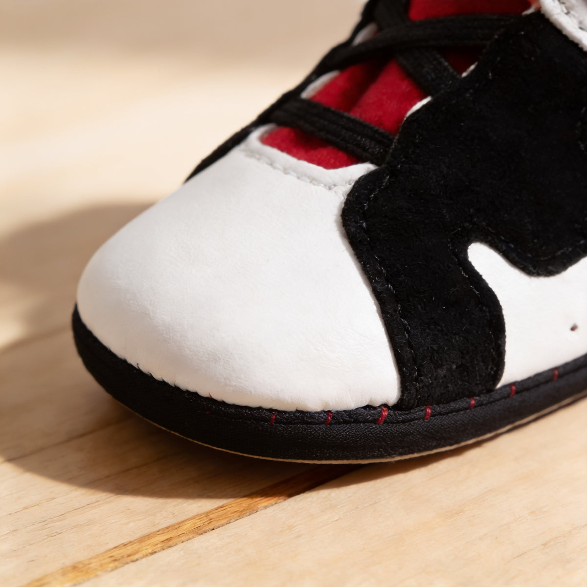 Close-up of a White & Black baby sneaker toe showing smooth white PU leather, black suede overlay, black elastic laces and the thin black cloth sole edge with red stitching on a pale oak floor