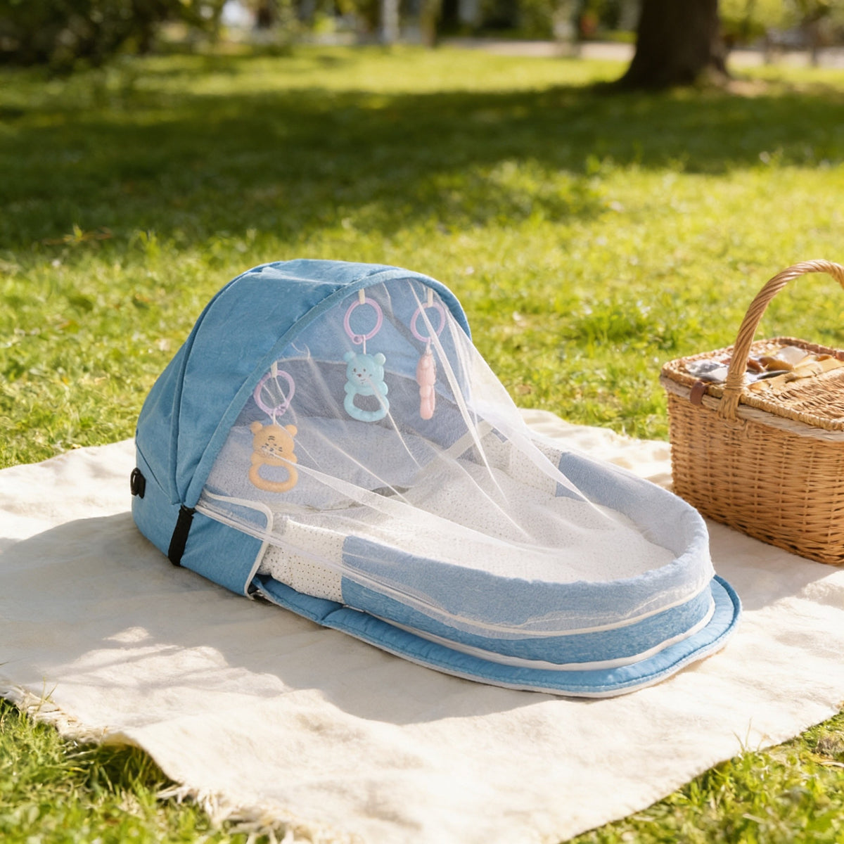 Blue portable travel bassinet with sunshade hood and mesh net on a picnic blanket in a park.