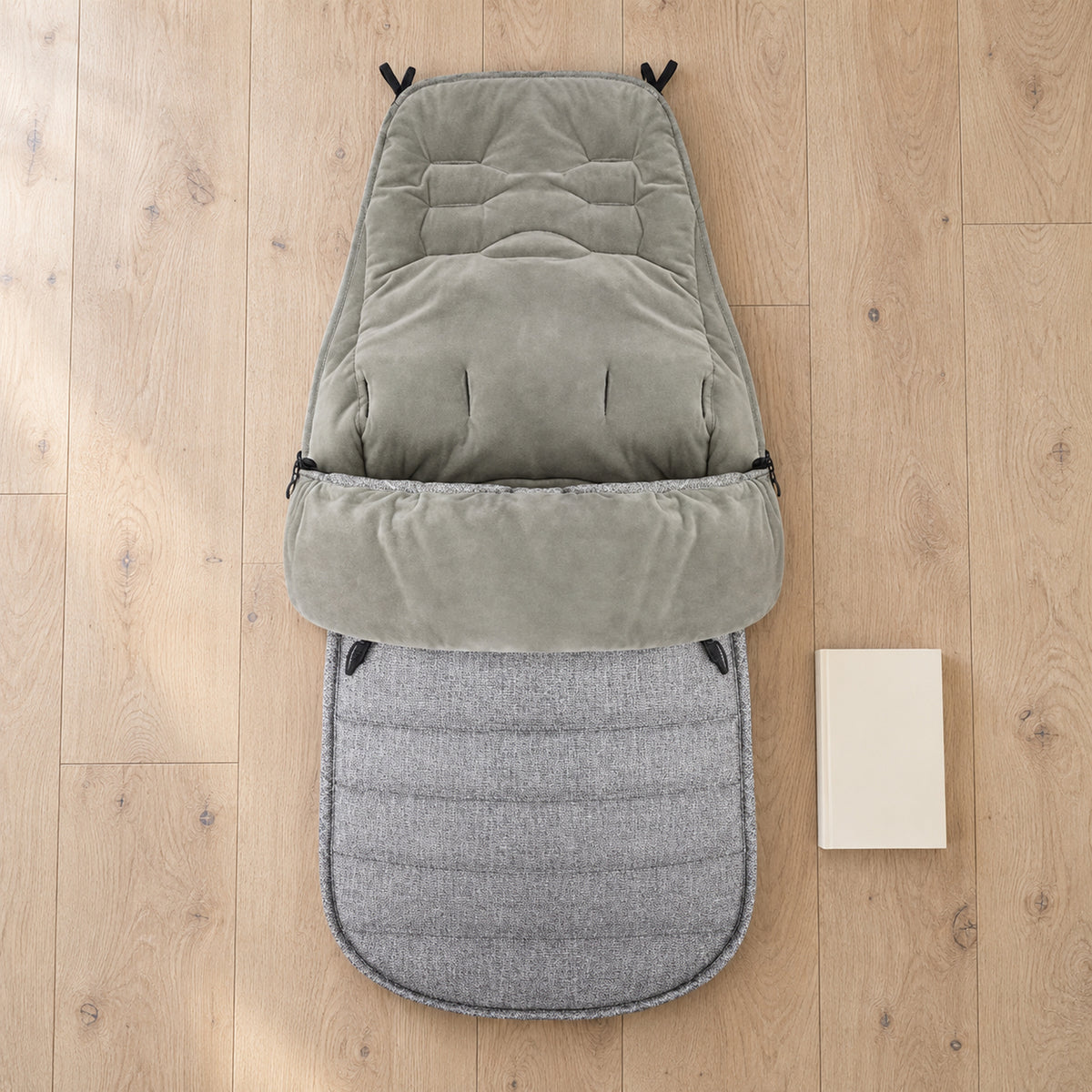 Gray winter stroller footmuff laid flat beside a plain book on a pale oak floor