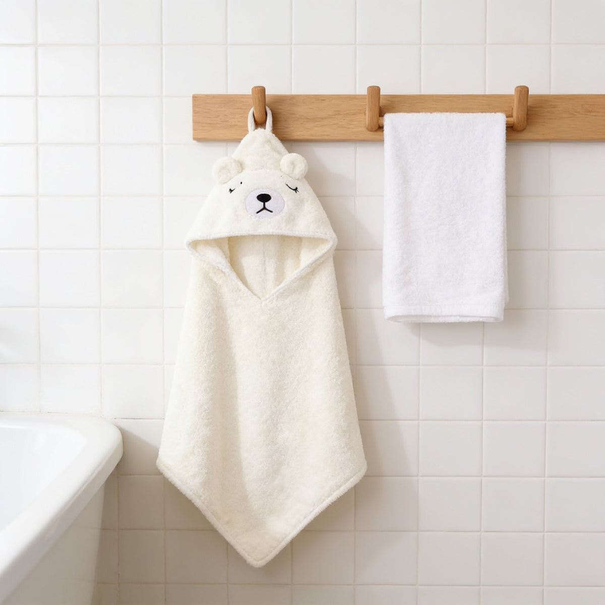 Cream bear hooded baby towel hanging from an oak peg rail in a white tiled bathroom.