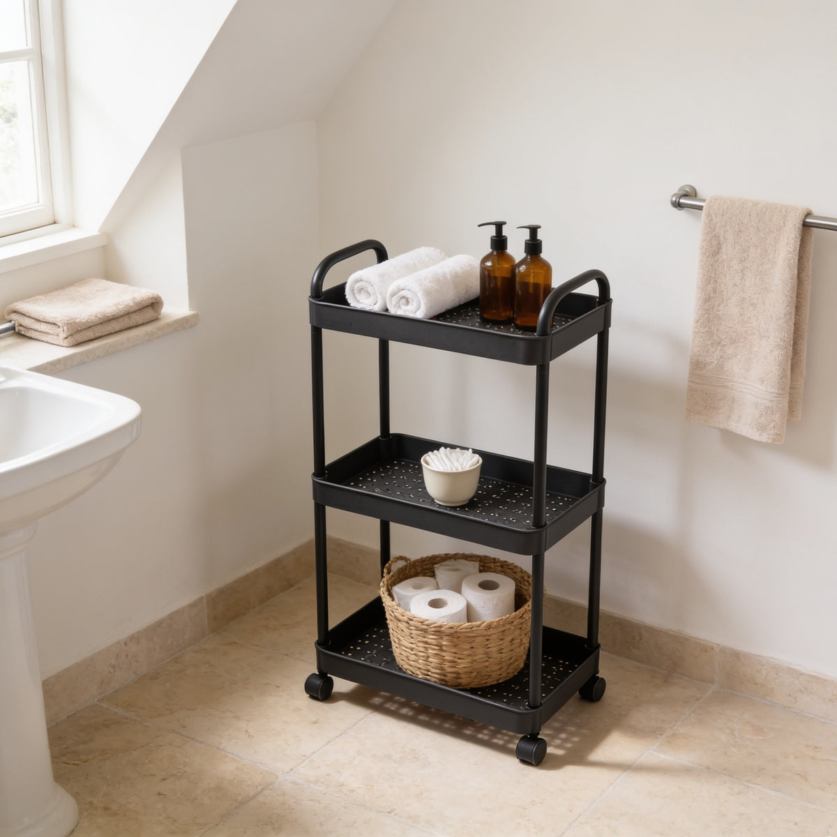 Black three-tier rolling utility cart beside a bathroom basin holding towels, bottles and spare rolls.
