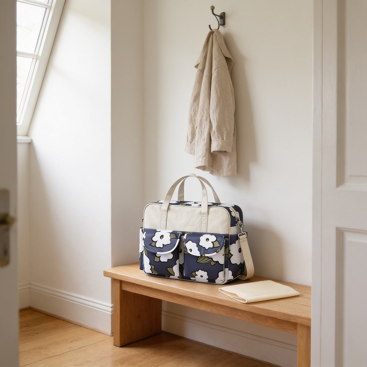 Navy floral print diaper tote bag with cream top band on a light oak hallway bench, its changing mat folded beside it.