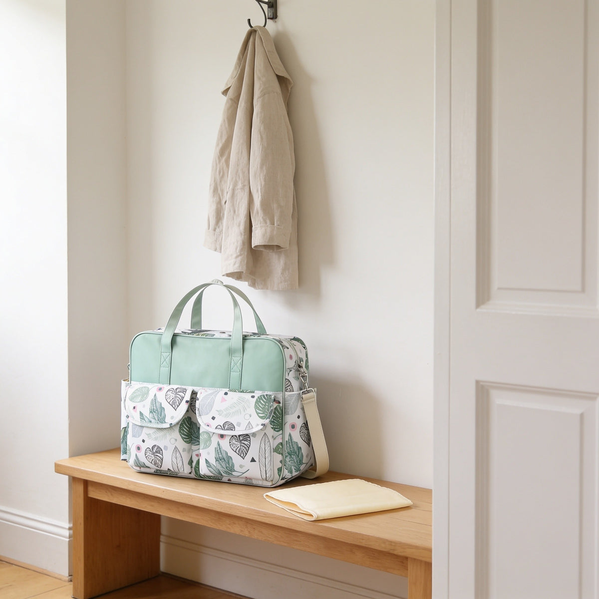 Mint botanical leaf print diaper tote bag on a light oak hallway bench, its changing mat folded beside it.