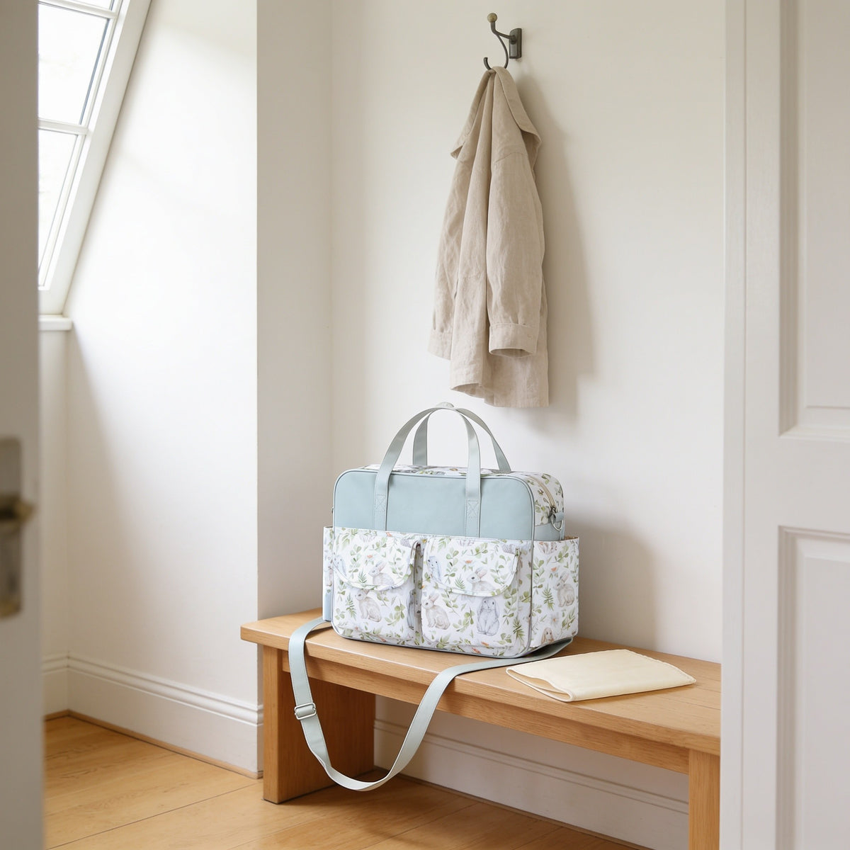 Rabbit meadow print diaper tote bag on a light oak hallway bench, its changing mat folded beside it.