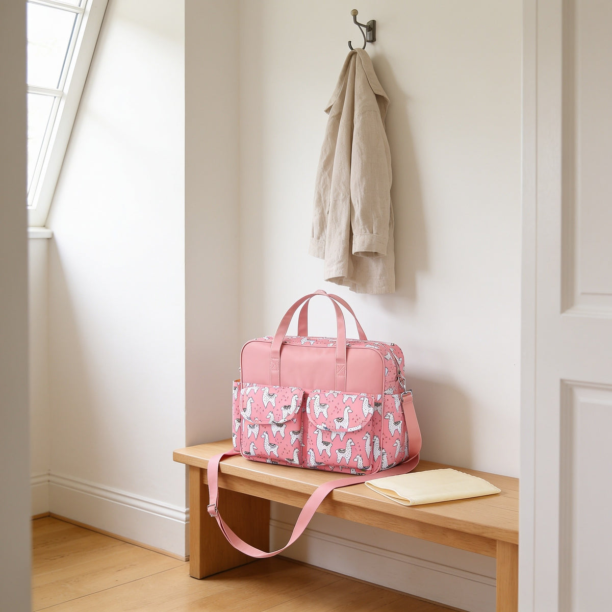 Pink alpaca print diaper tote bag on a light oak hallway bench, its changing mat folded beside it.
