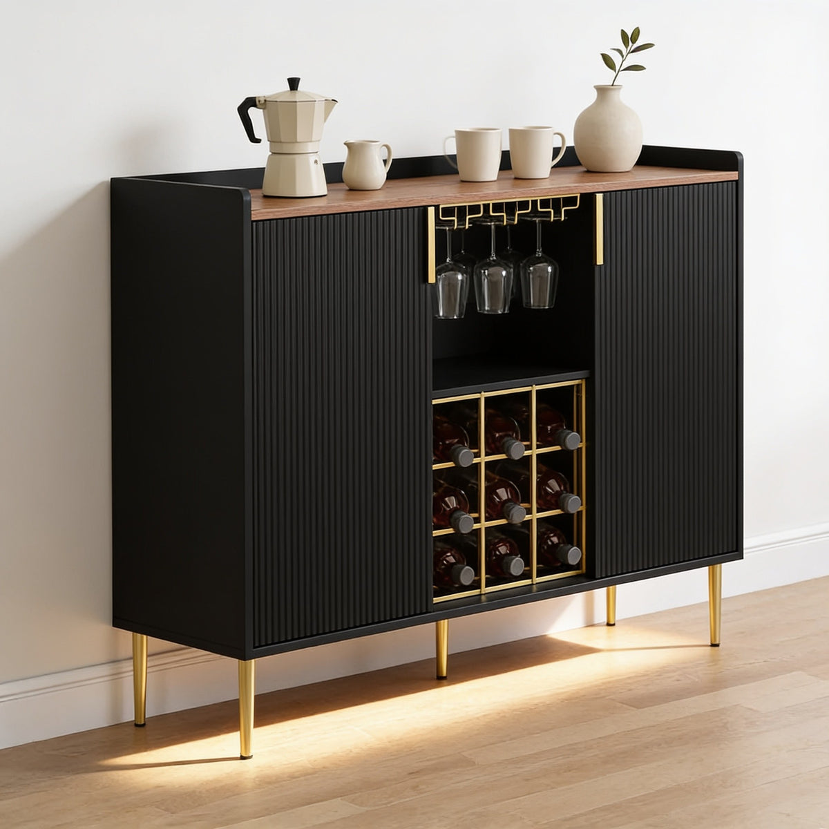 Black fluted coffee bar cabinet with a walnut top, brushed gold handles, a gold stemware rail and a nine-bottle gold wine rack, standing on gold legs against a bone-white wall.