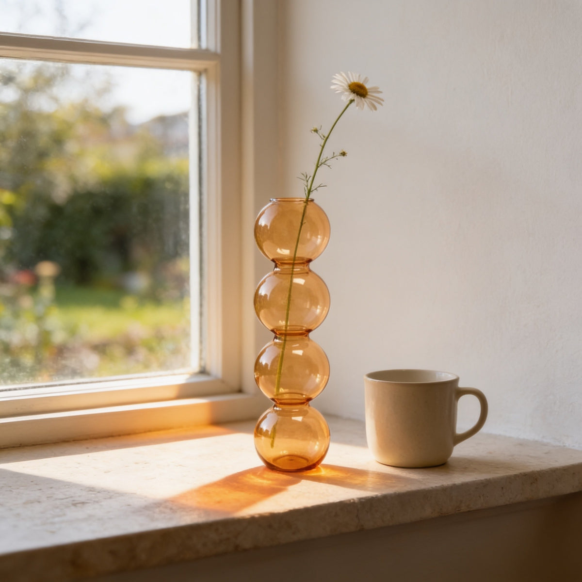 Amber stacked glass bubble bud vase with four bubbles holding a chamomile stem on a sunny windowsill beside a mug.