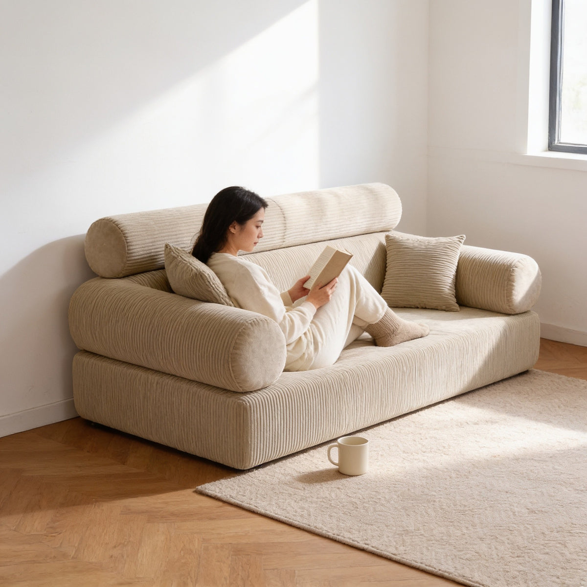 Adult sitting curled up on the beige corduroy folding sofa bed, reading, showing its sixty-five centimetre seat depth.