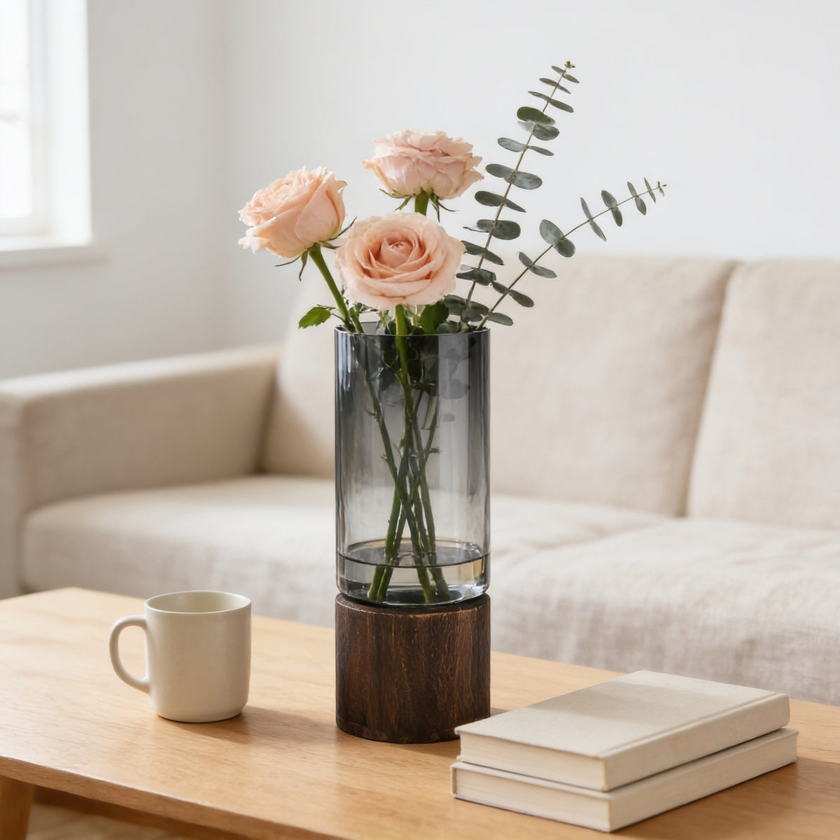 Smoked gray glass cylinder vase in a round walnut wood base holding peach roses on an oak coffee table.