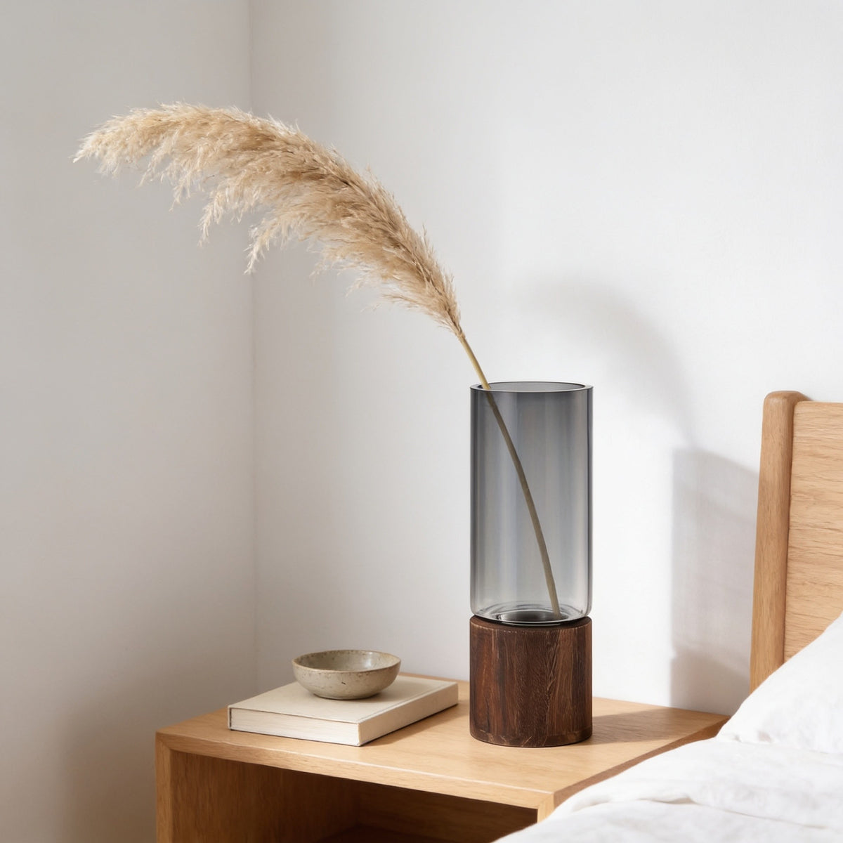 Smoked glass vase with a single stem of pampas grass on a bedside table.