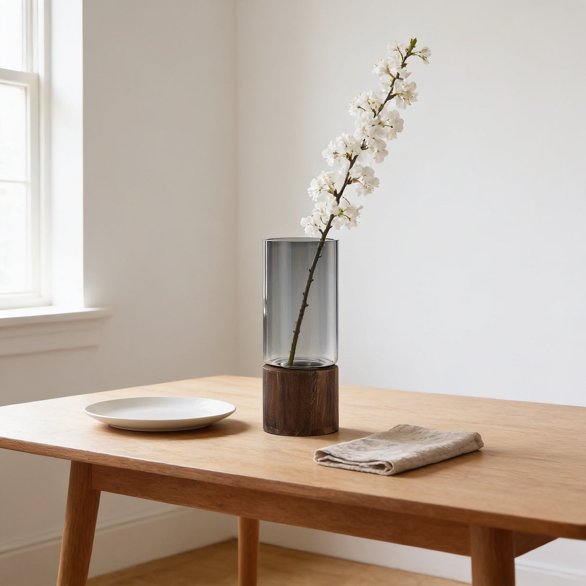 Smoked glass vase with a branch of white blossom on a dining table beside a dinner plate.