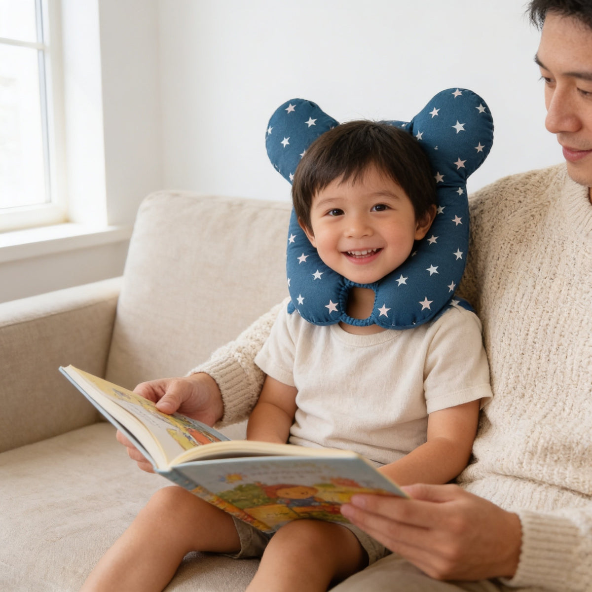 Toddler sitting awake beside a parent on a sofa, reading, wearing the navy star U-shaped cotton neck pillow.