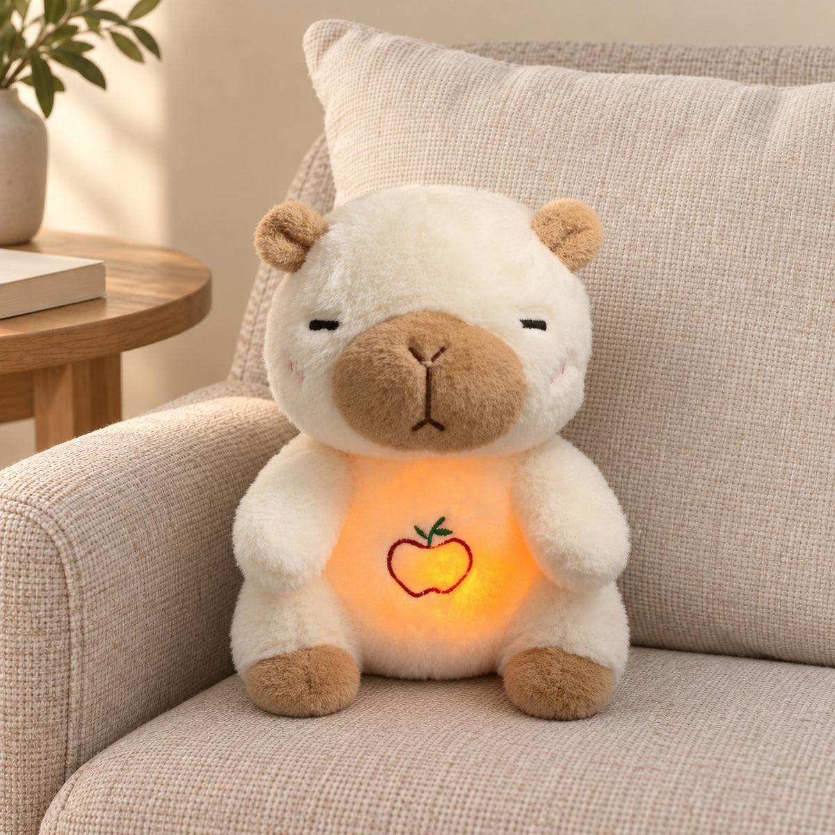 Ivory capybara plush with tan muzzle and glowing embroidered apple on a linen armchair