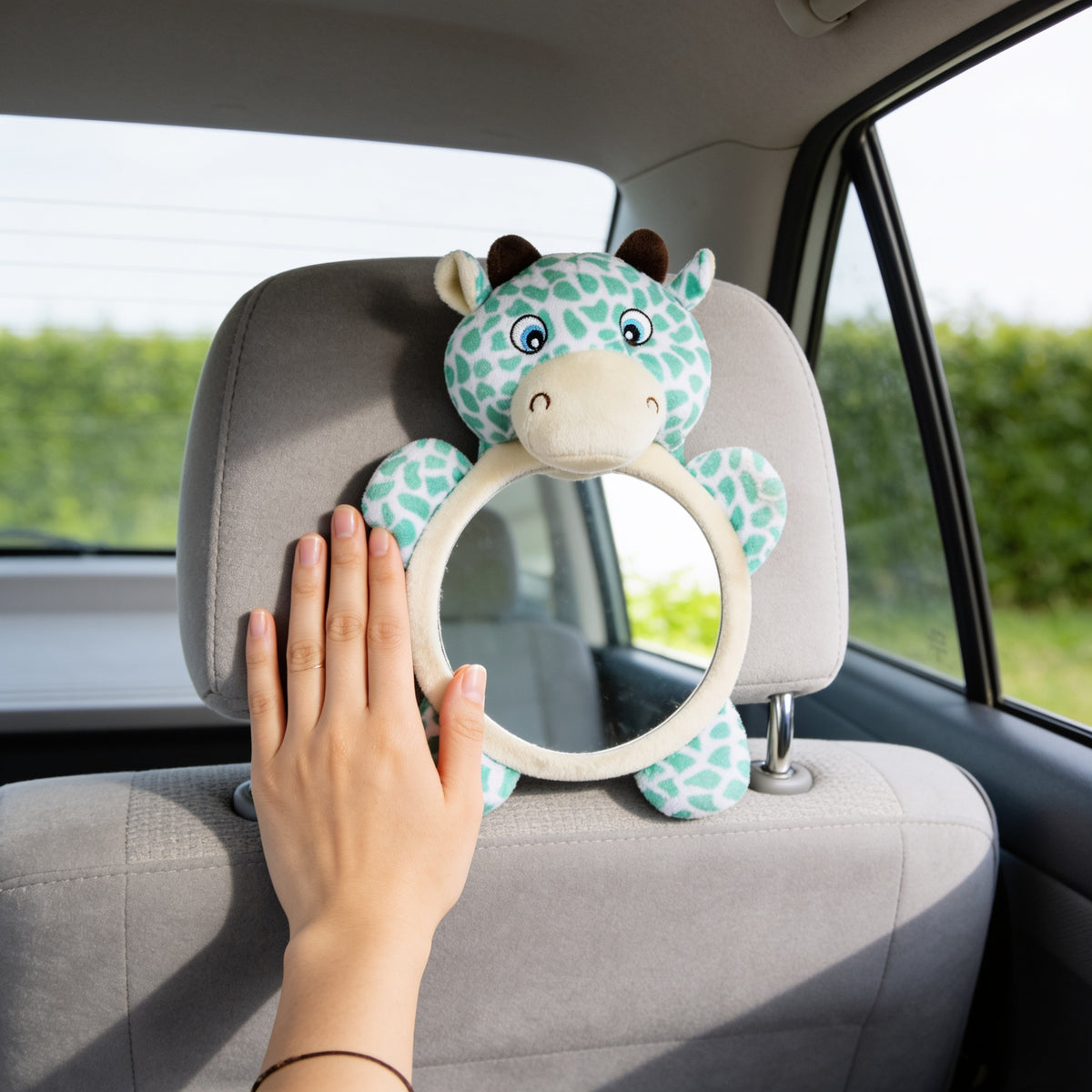Adult hand laid flat beside the Deer baby car mirror on a grey back-seat headrest, showing the size of the round mirror next to a hand