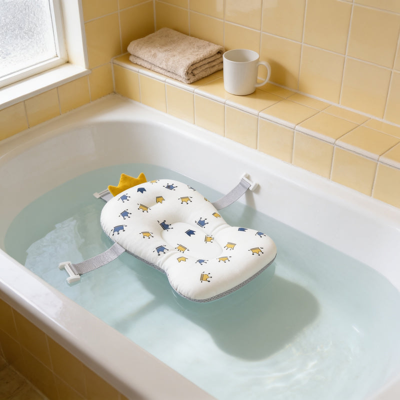 White floating baby bath pad printed with small navy and mustard crowns, with a mustard fabric crown at the head, floating empty on shallow clear water in a white bathtub beside a cream-tiled ledge with a folded oatmeal towel and a white mug