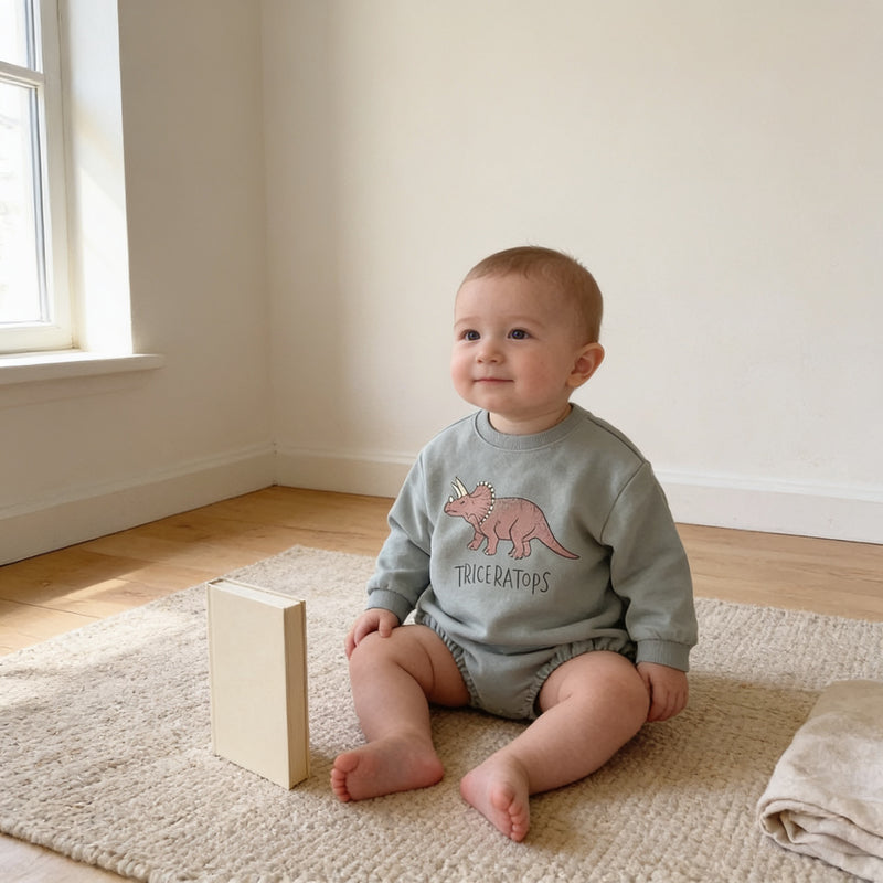 Sage-grey Triceratops long-sleeve bubble bodysuit worn by a smiling seated baby on an oatmeal flat-weave rug in a cream nursery, a plain cream book beside the baby