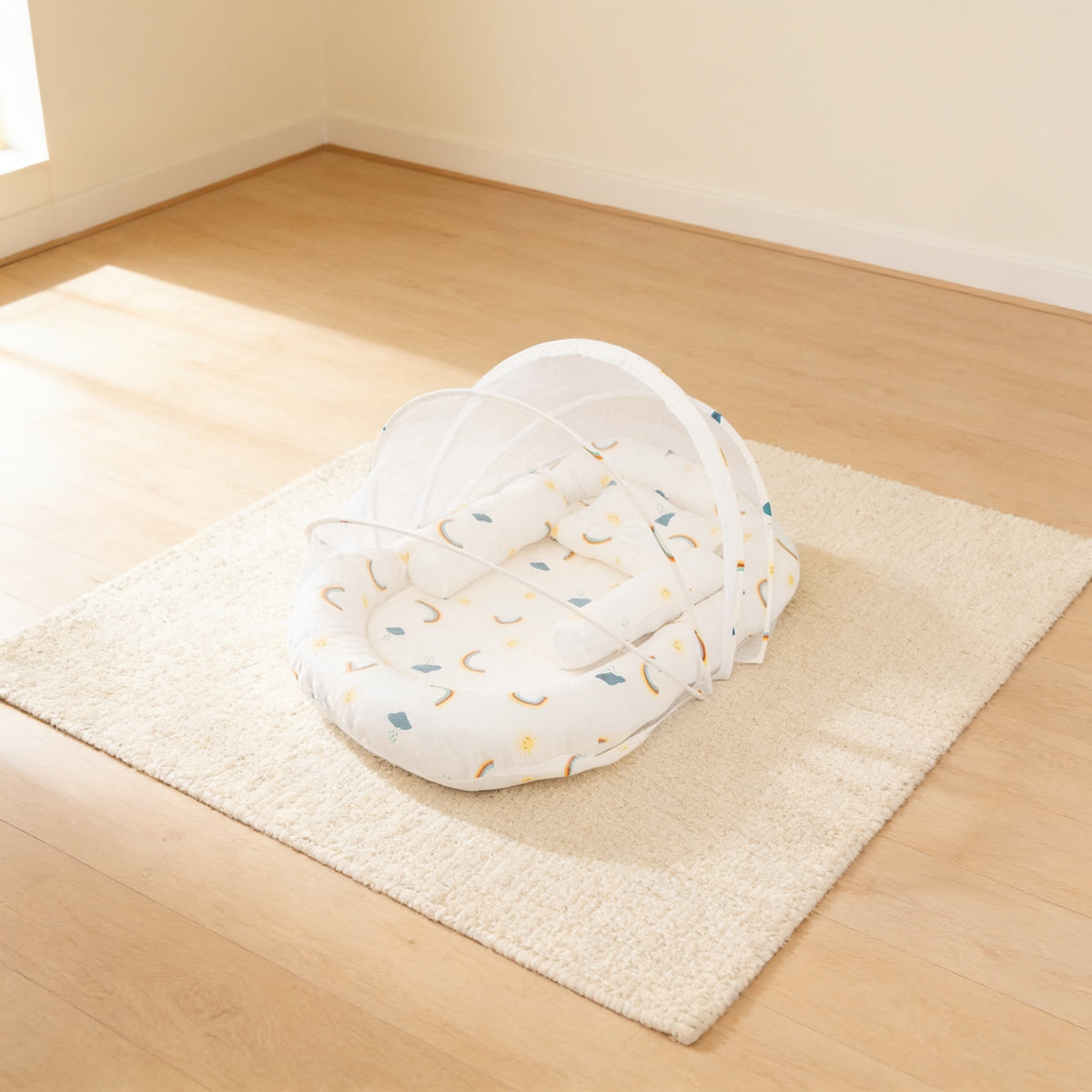 Cream Rainbows foldable travel bassinet, cream cotton with small pastel rainbows, with its mesh net dome up on a cream rug in a bright living room