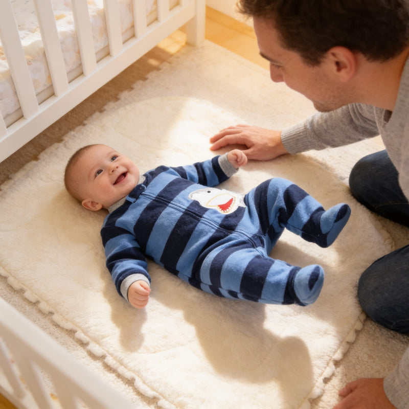 Awake smiling baby in blue shark stripe fleece footie pajamas on a play mat with a parent beside.