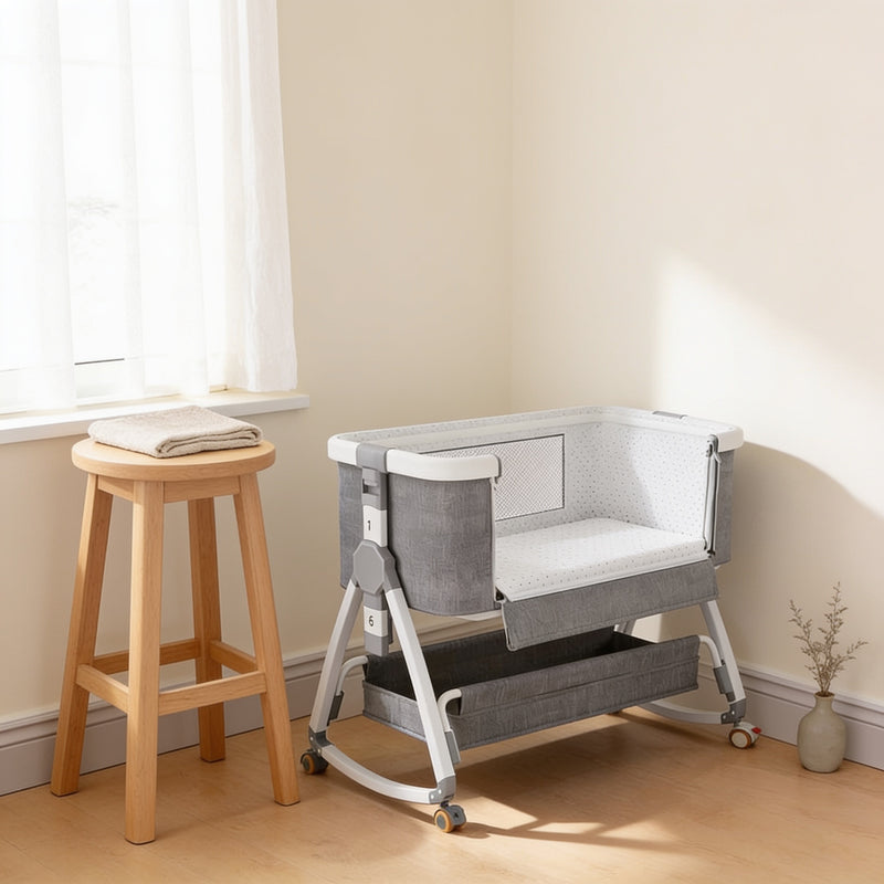 Grey and white height-adjustable rocking bedside bassinet standing on a pale oak floor in a cream nursery corner, near long side folded down to show the white dotted mattress inside, charcoal storage tray slung in the white aluminium frame below