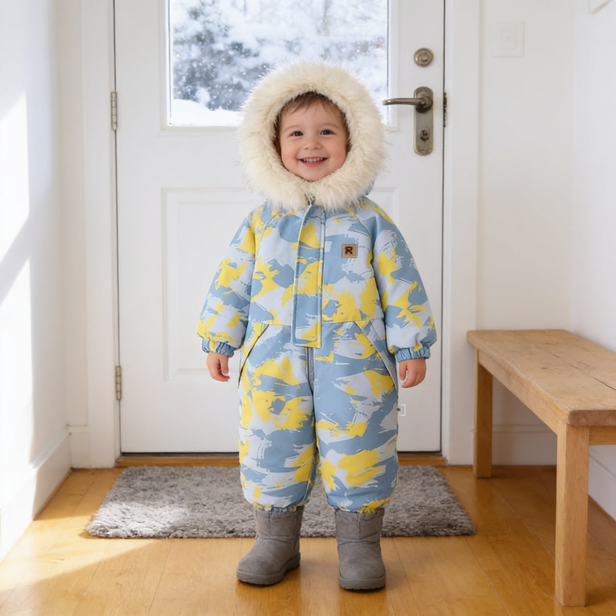Toddler wearing the Blue Camo hooded one-piece snowsuit with a cream fluffy hood trim, standing in a bright home entryway by a snowy front door