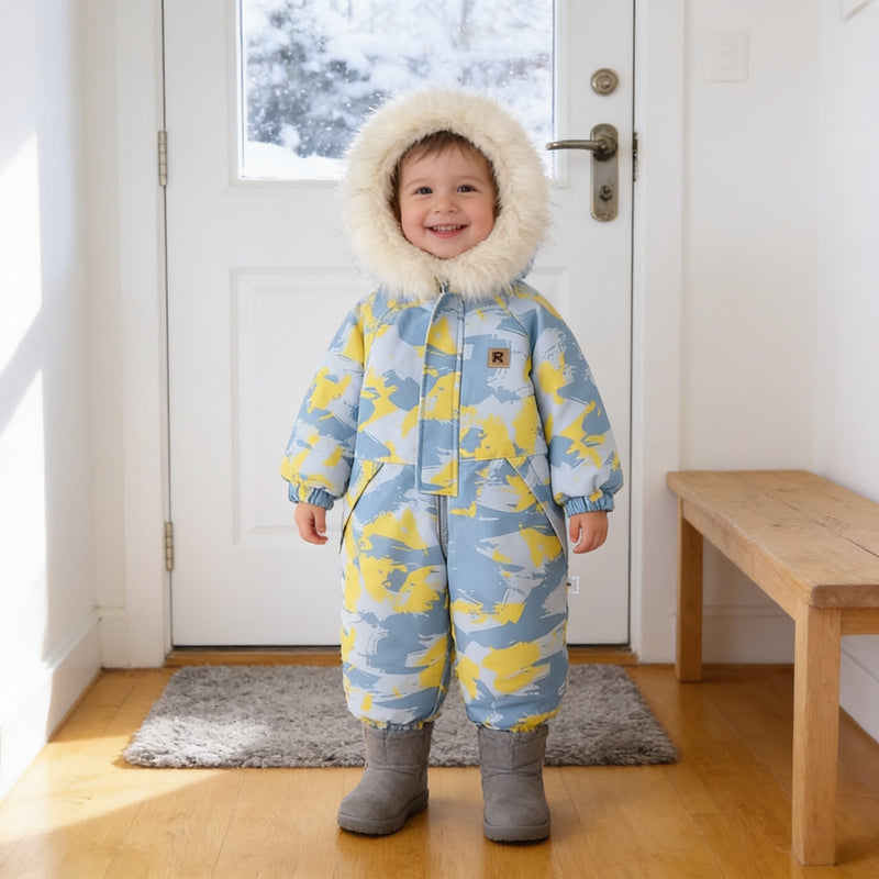 Toddler wearing the Blue Camo hooded one-piece snowsuit with a cream fluffy hood trim, standing in a bright home entryway by a snowy front door
