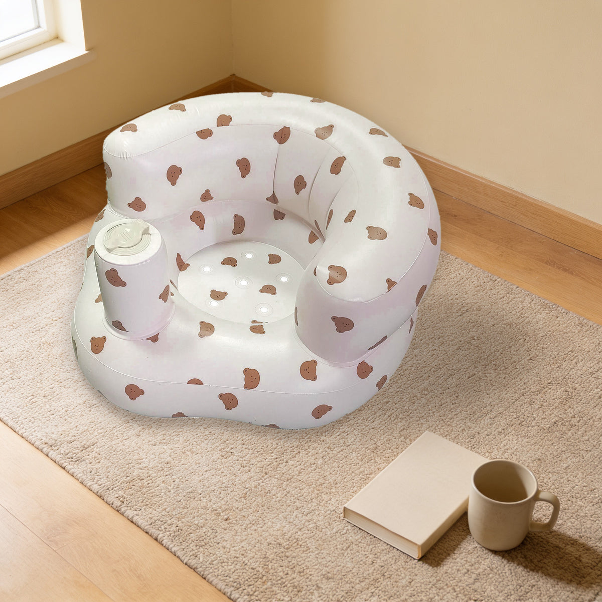 Bear design inflatable baby floor seat, milky off-white printed with small brown bear heads, empty on the same oatmeal woven rug beside a plain cream book and a stoneware mug in the warm cream living room