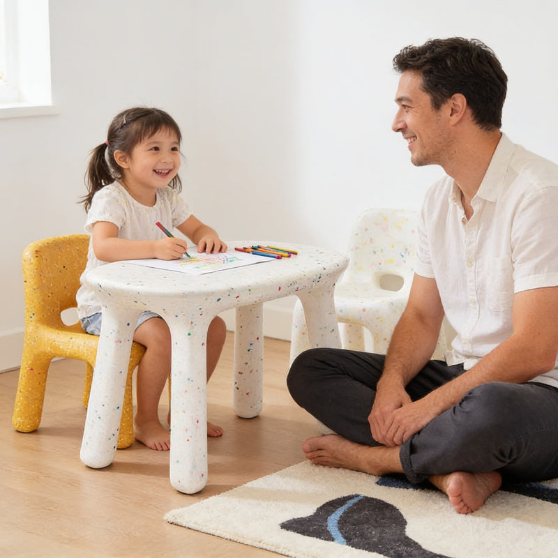 Girl drawing at the white speckled kids table on a yellow chair while her father sits on the rug beside her.