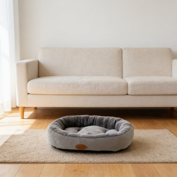 Gray oval plush cat and dog bed on an oatmeal rug in front of a linen sofa