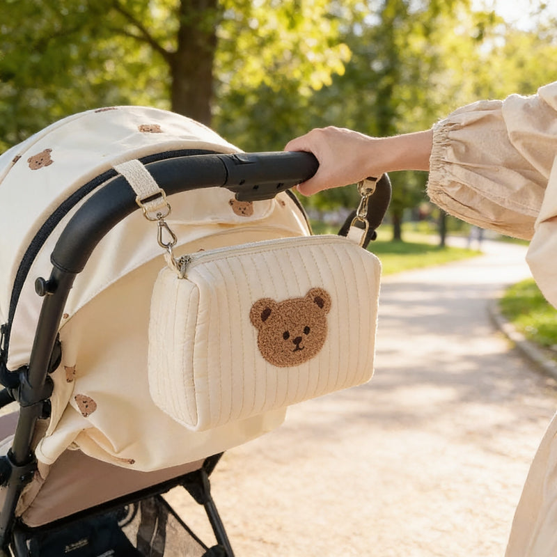 Cream quilted cotton diaper pouch with bear patch clipped to a stroller handlebar on a park walk.