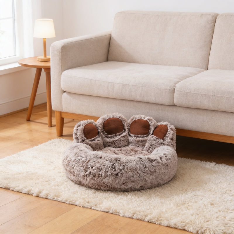 Brown paw-shaped plush dog and cat bed with four raised toes on a cream rug beside a linen sofa in a sunny living room