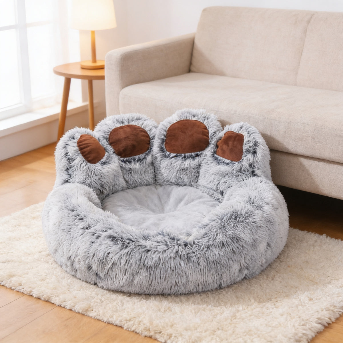 Gray paw-shaped plush dog and cat bed with brown toe pads on a cream rug beside a linen sofa