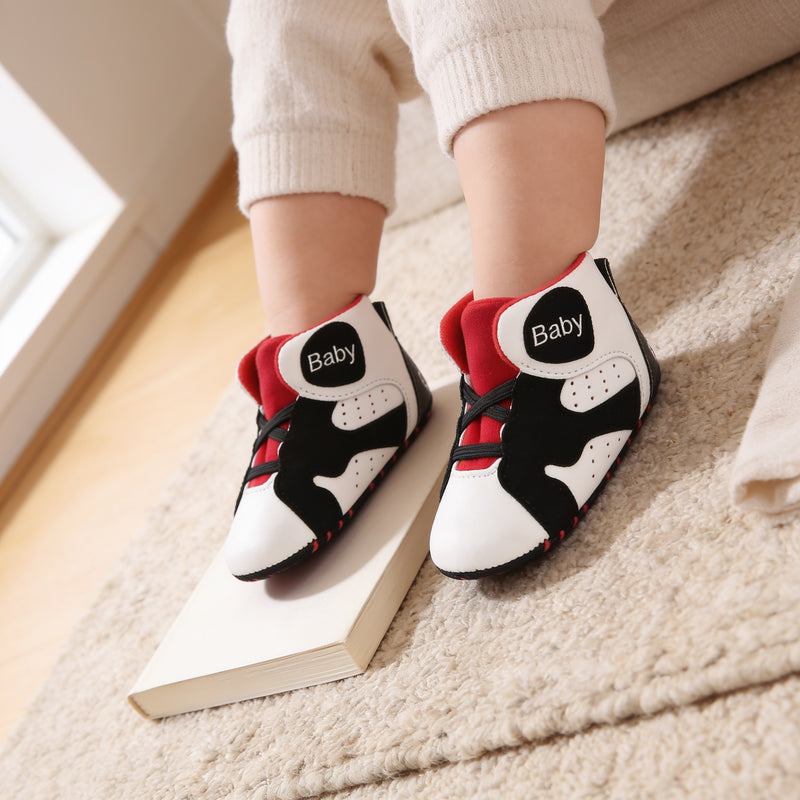 White & Black soft-sole baby high-top sneakers with red lining, black elastic laces and a black 'Baby' ankle patch, worn on a baby's feet cropped below the knee, resting on an oatmeal wool rug and a blank paperback in a sunlit cream living room