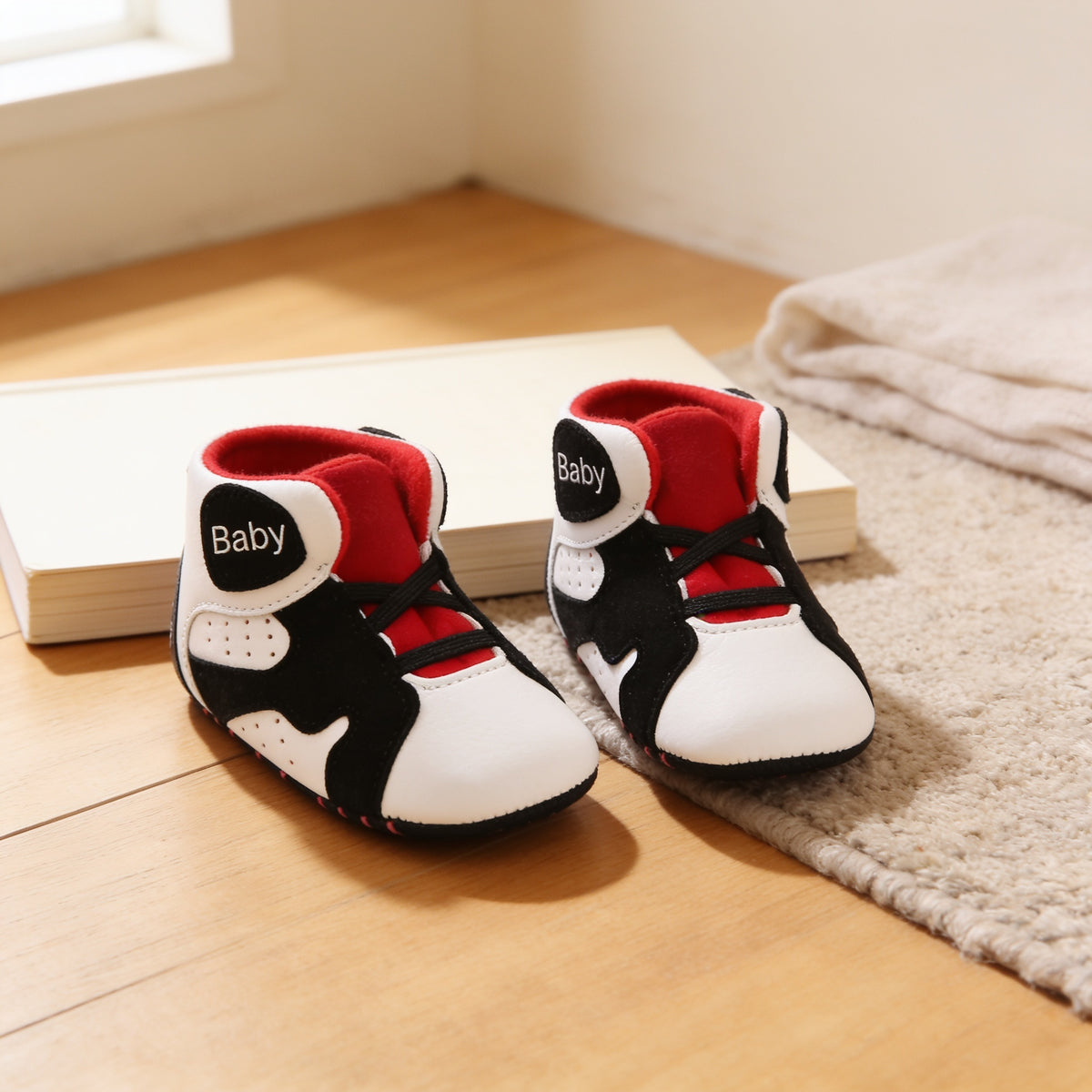 Pair of White & Black soft-sole baby high-top sneakers with red lining and black 'Baby' patches set on pale oak floorboards in front of a blank paperback, beside an oatmeal rug and folded muslin blanket