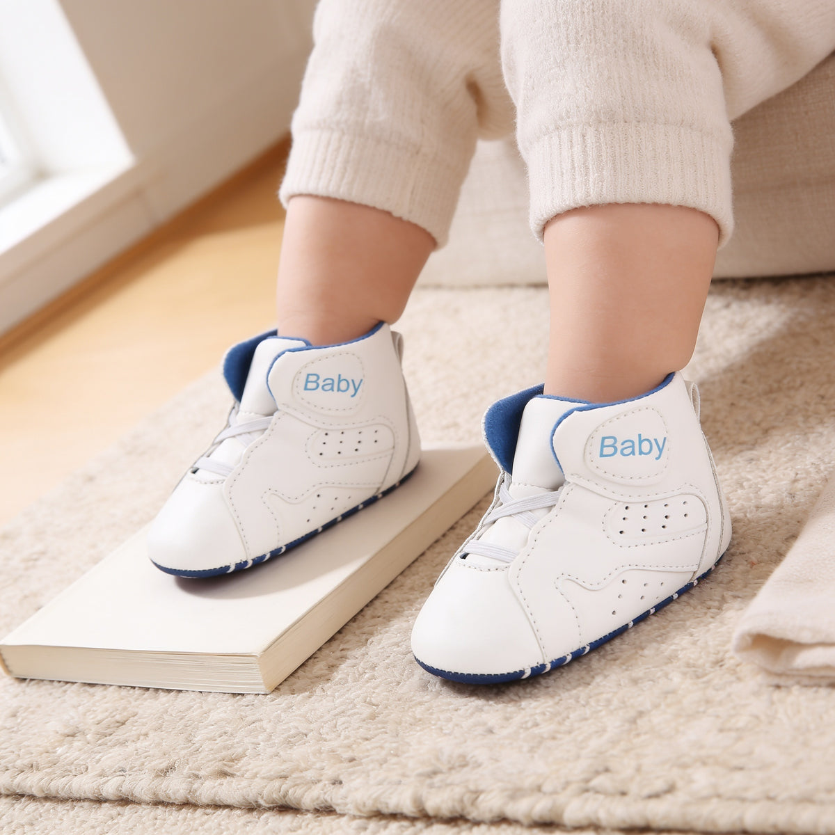 White soft-sole baby high-top sneakers with royal blue lining and sole edge, white elastic laces and a light-blue 'Baby' patch, worn on a baby's feet on an oatmeal rug and blank paperback in a sunlit living room