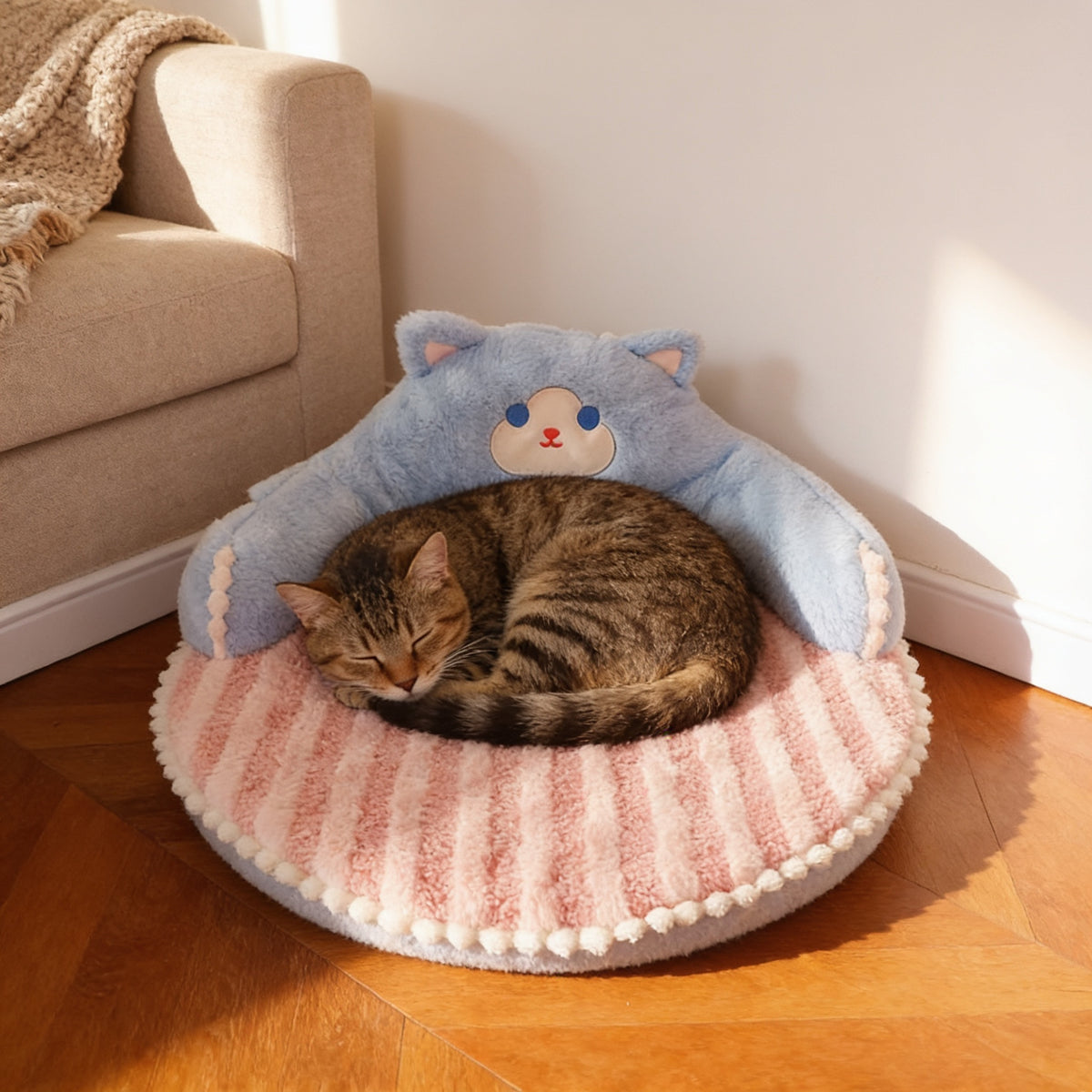 Tabby cat asleep on the round cat bed with a light-blue cat-shaped backrest and pink striped mat in a cosy living-room corner