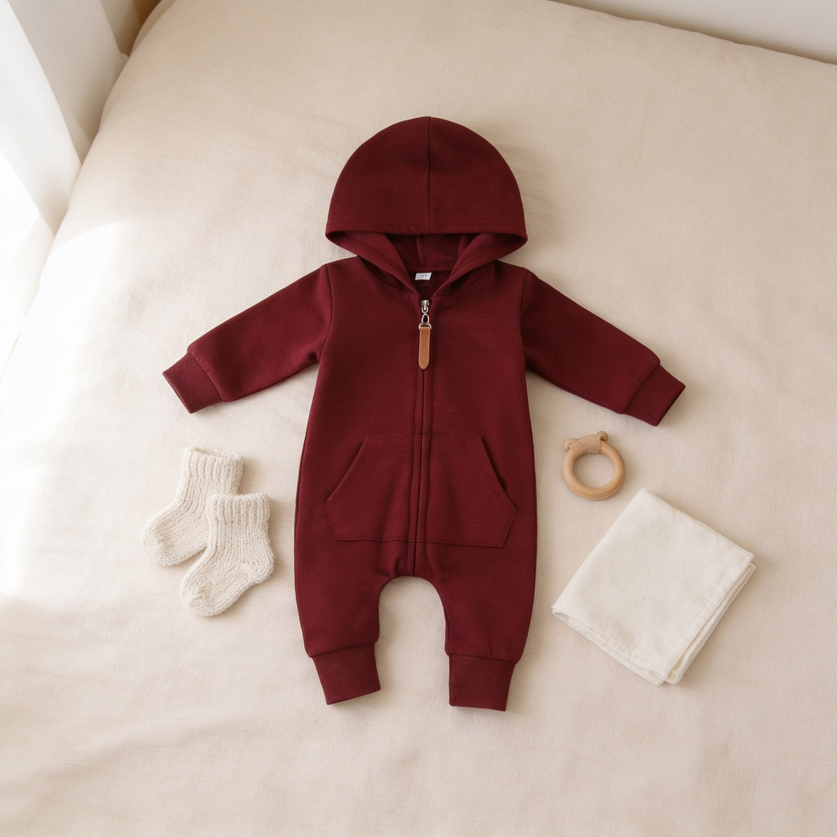 Burgundy zip-up hooded baby romper laid flat on a linen bedspread.